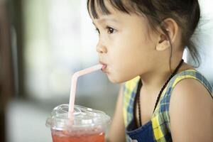 Porträt von ein wenig asiatisch Mädchen Trinken Süss Wasser im ein Glas mit ein Stroh. foto