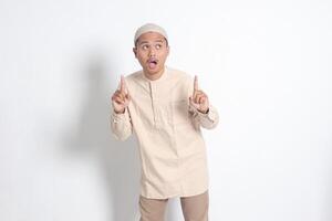 Porträt von schockiert asiatisch Muslim Mann im koko Hemd mit Schädeldecke zeigen Produkt und zeigen mit seine Hand und Finger hoch. Werbung Konzept. isoliert Bild auf Weiß Hintergrund foto