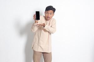 Porträt von jung aufgeregt asiatisch Muslim Mann im koko Hemd zeigen leer Bildschirm Handy, Mobiltelefon Telefon Attrappe, Lehrmodell, Simulation während zeigen und präsentieren Produkt. Sozial Medien Konzept. isoliert Bild auf Weiß Hintergrund foto