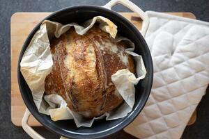 oben Aussicht von frisch hausgemacht Sauerteig Brot im Besetzung Eisen Pfanne, Ofen Handschuhe auf hölzern Schneiden Tafel foto