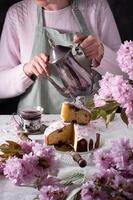 ein Frau gießt Tee von ein Silber Teekanne, Hintergrund von Rosa Sakura Blumen, Ostern foto