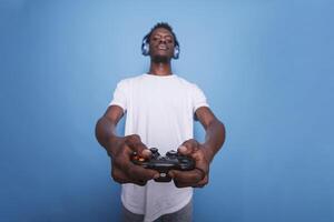 afrikanisch amerikanisch Video Spieler tragen Kopfhörer und halten ein Joystick zu das Kamera. jugendlich schwarz Mann Stehen im Vorderseite von Blau Hintergrund während mit ein Gerät zum online Spielen. foto