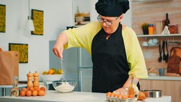 Bäcker Verbreitung Mehl auf hölzern Tabelle beim Zuhause im modern Küche tragen Schürze und Bonete. glücklich Alten Koch mit Uniform streuen, Sieben Sichtung roh Zutaten durch Hand Backen hausgemacht Pizza foto