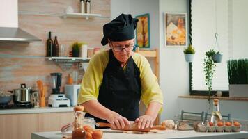 im Ruhestand Frau Koch mit hölzern rollen Stift zum Kochen Pizza. glücklich Alten Bäcker mit Bonete vorbereiten roh Zutaten zu Backen traditionell Pizza streuen, Sieben Mehl auf Tabelle im das Küche. foto