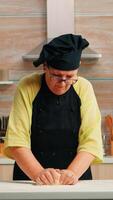 Oma vorbereiten Weizen hausgemacht Brot tragen Koch Bonete. im Ruhestand Alten Bäcker mit Bonete Mischen Zutaten mit Weizen Mehl kneten Teig zum Backen hausgemacht Kuchen und Brot foto