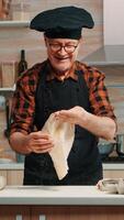 Koch werfen oben Teig zum Pizza beim Zuhause im modern Küche lächeln im Vorderseite von Kamera. geschickt im Ruhestand Alten Koch tragen Uniform Spinnen und werfen Pizza Arbeitsplatte foto