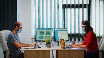 Menschen mit Maske und Visier Arbeiten im Neu normal Büro Überprüfung Berichte und Schreiben auf Computer. Mitarbeiter im modern Arbeitsplatz respektieren Schutz Regeln gegen covid Virus mit Plexiglas. foto