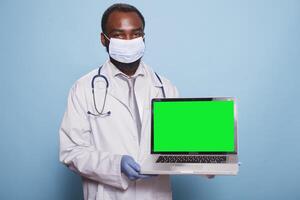 Arzt tragen medizinisch schützend Ausrüstung präsentieren Laptop mit Grün Bildschirm Attrappe, Lehrmodell, Simulation zum Gesundheitswesen Konzept. afrikanisch amerikanisch Arzt halten Minicomputer Anzeigen leer Chromakey Vorlage. foto