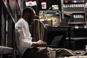 Privat Ermittler Spionage, Aufpassen cctv Kamera Aufzeichnungen auf Laptop während Sitzung auf Fußboden im dunkel Agentur Büro. afrikanisch amerikanisch Detektiv Analysieren Verbrechen Fall Fotos auf Computer