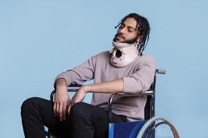arabisch Mann tragen zervikal Halsband und Schlafen während Sitzung im Rollstuhl. jung erschöpft geduldig mit geschlossen Augen erholen von Unfall, mit Hals Klammer zum Wirbelsäule Schutz foto