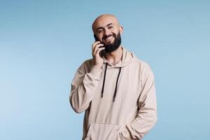 heiter lächelnd arabisch Mann reden auf Smartphone Porträt. glücklich jung Person antworten Forderung, chatten auf Handy, Mobiltelefon Telefon während Stehen und suchen beim Kamera mit sorglos Ausdruck foto