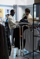 Kleidung Geschäft Mitarbeiter assistieren Kunde im Auswahl modisch Kleidung, Angebot Orientierungshilfe auf Stil. Boutique Arbeiter präsentieren Jacke zu kaukasisch Klient und geben Rat foto