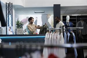 afrikanisch amerikanisch Kunde Stehen beim Kleidung Geschäft Zähler während Kassierer Scannen Kleidung. Mann warten beim Kasse registrieren Schreibtisch und Einkauf Kleidungsstück während Einkaufen im Boutique foto