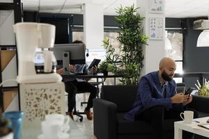 arabisch Geschäft Arbeiter chatten mit Mitarbeiter mit Smartphone während Arbeiten im modern öffnen Raum. Marketing Fachmann Sitzung auf Couch und SMS im Sozial Medien auf Handy, Mobiltelefon Telefon im Start oben Büro foto