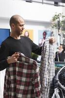Einkaufen Einkaufszentrum Boutique arabisch Mann Kunde Surfen Plaid Hemden und halten zwei Outfits auf Kleiderbügel. Kleidung Geschäft Klient wählen beiläufig Herrenbekleidung Kleidung, in Anbetracht Stil und passen foto