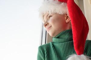 Kinder- Träume Konzept. Freundschaft. ein einsam wenig Junge im ein Santa claus Hut ist traurig beim das Fenster warten zum Geschenke, suchen aus das Fenster, das Kind ist warten zum Santa Klaus. foto