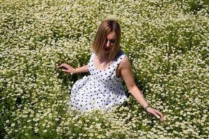 ein Frau im ein Weiß Kleid mit Polka Punkte ist auf ein Lichtung mit Gänseblümchen. Blühen Gänseblümchen foto