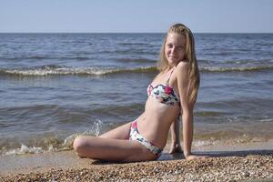 das Mädchen das blond im ein Bikini Sitzung auf das Strand im das Sand. schön jung Frau im ein bunt Bikini auf Meer Hintergrund foto