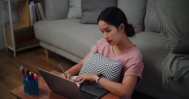 Porträt von jung Frau Sitzung auf das Fußboden gelehnt gegen ein Sofa Arbeiten mit ein Laptop und nehmen Anmerkungen im ein Notizbuch beim Zuhause Büro. foto