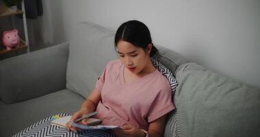 Porträt von jung Frau Sitzung auf Sofa suchen durch Arbeitsblatt und Überprüfung Daten und nimmt Anmerkungen beim Zuhause Büro foto