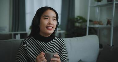 Porträt von glücklich jung asiatisch Frau genießen Hören zu das Musik- mit kabellos Kopfhörer während Sitzung trinken Kaffee auf Sofa im Leben Zimmer beim heim, Freizeit und Lebensstil, kostenlos Zeit foto