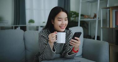Porträt von glücklich jung asiatisch Frau sitzen auf Sofa genießen chatten auf Handy, Mobiltelefon Telefon während Trinken Kaffee im Morgen im Leben Zimmer, frei Zeit, nimm Pause, lächelnd foto