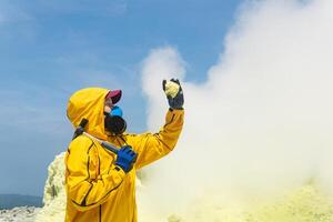 Frau Vulkanologe auf das Hintergrund von ein Rauchen Fumarole untersucht ein Stichprobe von ein Schwefel Mineral foto