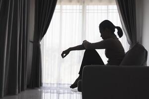 Silhouette von ein Person Leiden von Depression im das Haus, deprimiert Frau Sitzung allein auf das Sofa Gefühl betonen, traurig und besorgt im das dunkel Zimmer. Person sind betont. foto