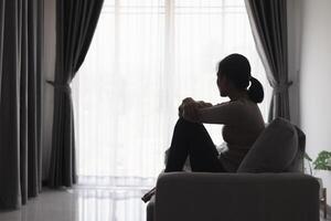 Silhouette von ein Person Leiden von Depression im das Haus, deprimiert Frau Sitzung allein auf das Sofa Gefühl betonen, traurig und besorgt im das dunkel Zimmer. Person sind betont. foto