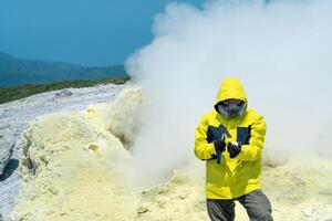 Mann Vulkan Wissenschaftler auf das Hintergrund von ein Rauchen Fumarole untersucht ein Stichprobe von ein Schwefel Mineral mit ein geologisch Hammer foto
