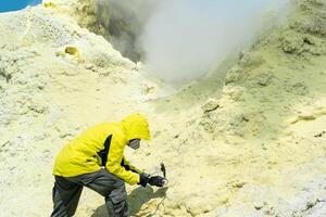 männlich Vulkanologe auf das Steigung von ein Vulkan Nächster zu ein Rauchen Schwefel Fumarole untersucht ein Stichprobe von ein Mineral foto