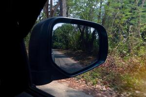 Aussicht von das Autos Seite Spiegel zu das Straße und Wald. fahren schnell Geschwindigkeit können sehen Grün Glas neben Straße. foto