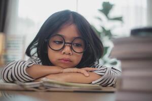 Kleines asiatisches Mädchen, das allein sitzt und mit gelangweiltem Gesicht herausschaut, Vorschulkind, das den Kopf auf den Tisch legt, mit trauriger Langeweile bei den Hausaufgaben, verwöhntes Kind foto