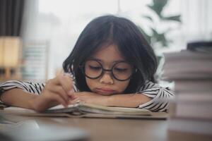 Kleines asiatisches Mädchen, das allein sitzt und mit gelangweiltem Gesicht herausschaut, Vorschulkind, das den Kopf auf den Tisch legt, mit trauriger Langeweile bei den Hausaufgaben, verwöhntes Kind foto