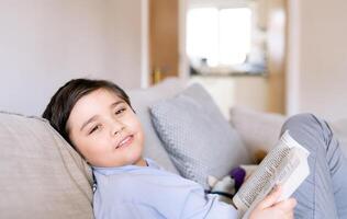 heiter Schule Kind Junge halten Buch Sitzung auf Sofa im Leben Zimmer, zurück zu Schule Konzept jung Kind lesen Buch oder tun Zuhause Arbeit beim Zuhause auf Wochenende, a Junge lächelnd und suchen beim Kamera foto