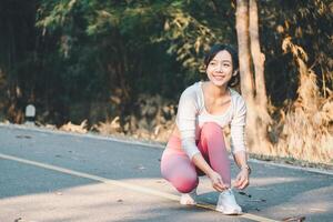 Fitness Konzept, strahlend jung Frau Schnürung ihr Schuhe mit ein lächeln, bekommen bereit zum ein Morgen Joggen im ein sonnendurchflutet bewaldet Park. foto