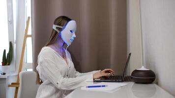 ein schön Mädchen mit ein LED Maske auf ihr Kopf funktioniert beim ein Laptop. Zuhause Haut Pflege Konzept. foto