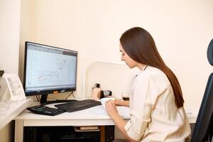 Porträt von ein glücklich weiblich Designer Schätzer Sitzung beim das Tabelle mit ein Notizbuch und Taschenrechner, Zählen das Balance und Tippen auf ein Laptop, Fertigstellung ihr Arbeit mit ein Tasse von Kaffee. foto