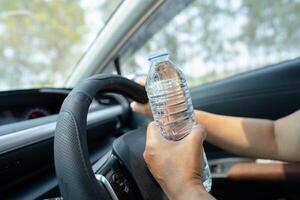 asiatische Fahrerin, die eine Flasche zum Trinken von Wasser hält, während sie ein Auto fährt. Wärmflasche aus Kunststoff verursacht Feuer. foto