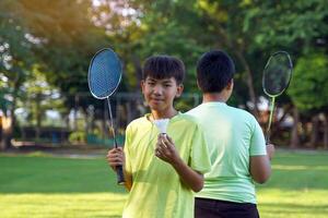 ein asiatisch Junge hält ein Badminton Schläger und ein Weiß Federball während spielen Badminton mit freunde auf das Park Rasen im das Abend nach Rückkehr von Schule. foto