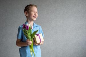 ein süß Junge ist halten ein bunt Strauß von Tulpen und ein Box mit ein Geschenk im seine Hände. foto