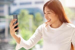 Menschen genießen mit Internet Video Berufung glücklich lächeln. asiatisch Frau suchen beim Smartphone lächelnd draussen. foto