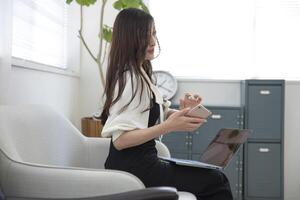 ein japanisch Frau Überprüfung Smartphone durch Fernbedienung Arbeit im das klein Büro foto