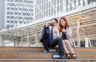 ein froh Geschäftsmann und Frau nehmen ein Selfie während Sitzung zusammen auf das Stufen, jung asiatisch Geschäft Menschen Mannschaft diskutieren im das Morgen foto