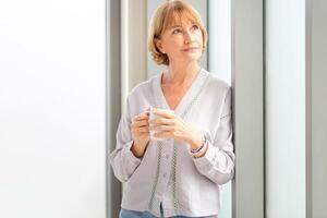 entspannend Frau Stehen in der Nähe von Fenster mit Tassen von Kaffee, Frau Innerhalb Neu Zuhause während ein Kaffee brechen foto