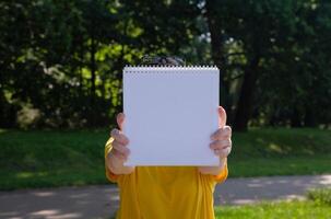 jung Frau im Gelb t Hemd halten ein Weiß Buch im Vorderseite von ihr im das Park auf ein sonnig Sommer- Tag. Hintergrund ist verschwommen. selektiv Fokus. hoch Qualität Foto