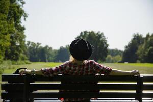 jung Frau im ein Hut und ein Plaid Hemd sitzt auf ein Bank in der Nähe von ein Wiese im das Park.Hintergrund verschwommen. selektiv Fokus. hoch Qualität Foto