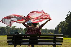 jung Mädchen sitzt auf ein Bank und hält ein Schal mit ein Muster von das amerikanisch Flagge im ihr Hände winken im das Wind. hoch Qualität Foto