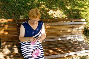 Senior Frau strickt im das Park auf ein Bank auf ein sonnig Tag. foto