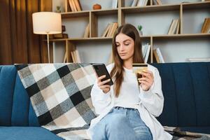 glücklich jung Frau halten Telefon und Anerkennung Karte Sitzung auf das Couch. online Einkaufen. foto